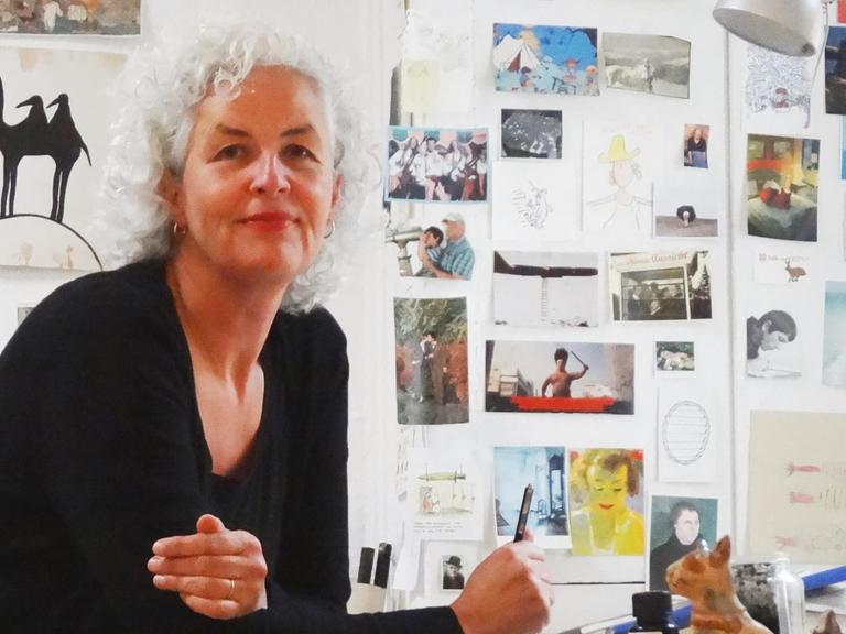 Die Cartoonistin Miriam Wurster sitzt an ihrem Zeichentisch. Um sie herum sind Postkarten, Skizzen und Pinsel zu sehen