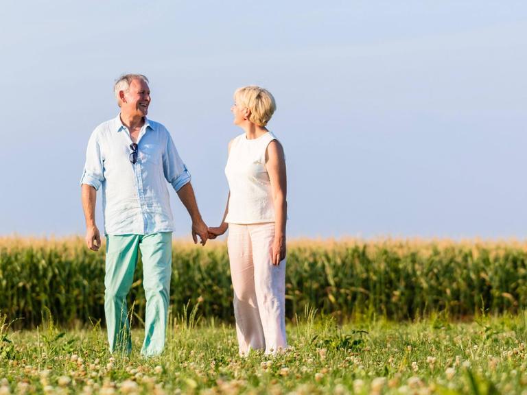 Älteres Paar steht glücklich lachend auf einer grünen Wiese vor blauem Himmel