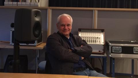 Ein Mann sitzt inmitten eines Studios mit verschränkten Armen und lächelt in die Kamera.