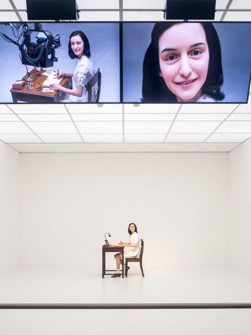 Die Videoinstallation "Likeness" (2018) von Simon Fujiwara im Hamburger Bahnhof in Berlin, die sich mit der medienwirksamen Inszenierung von Anne Frank auseinandersetzt. Eine Wachsskulptur von Anne Frank schreibt ihr berühmtes Buch und lächelt ununterbrochen, während eine Roboterkamera sie dabei filmt.