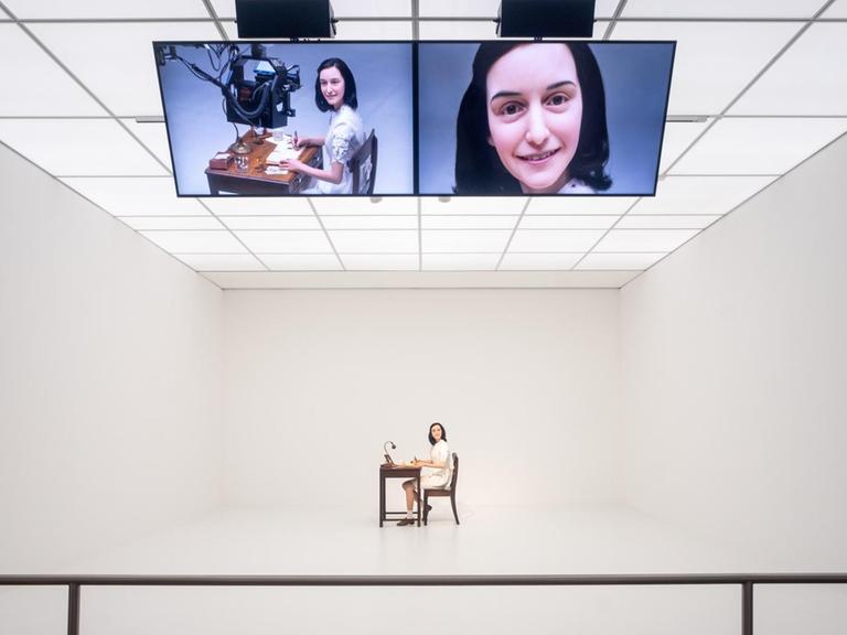 Die Videoinstallation "Likeness" (2018) von Simon Fujiwara im Hamburger Bahnhof in Berlin, die sich mit der medienwirksamen Inszenierung von Anne Frank auseinandersetzt. Eine Wachsskulptur von Anne Frank schreibt ihr berühmtes Buch und lächelt ununterbrochen, während eine Roboterkamera sie dabei filmt.