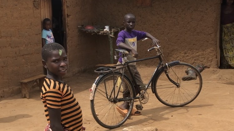 Drei afrikanische Kinder spielen in der Flüchtlingssiedlung Nakivale vor einem Steinhaus. Eines der Kinder stützt sich auf ein Fahrrad.