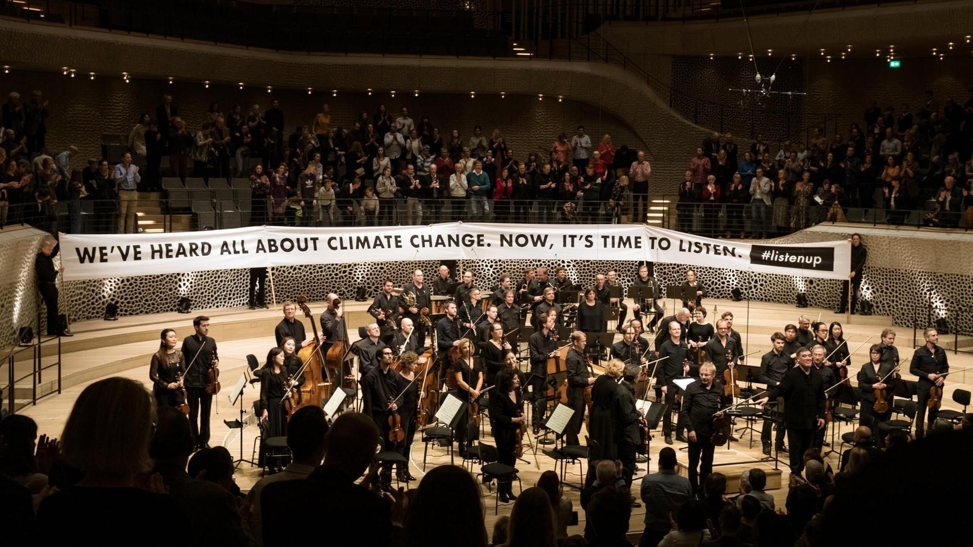 Ein Orchester steht vor einem Transparent, auf dem zu lesen ist "We've heard all about climate change, now it's time to listen"