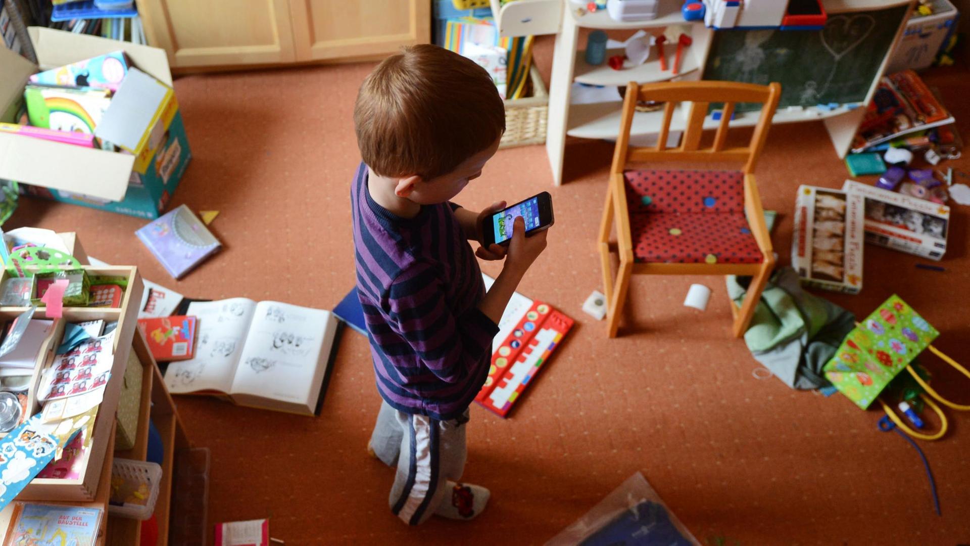 Ein fünfjähriger Junge spielt in seinem unaufgeräumten Kinderzimmer auf einem iPhone ein Computerspiel.