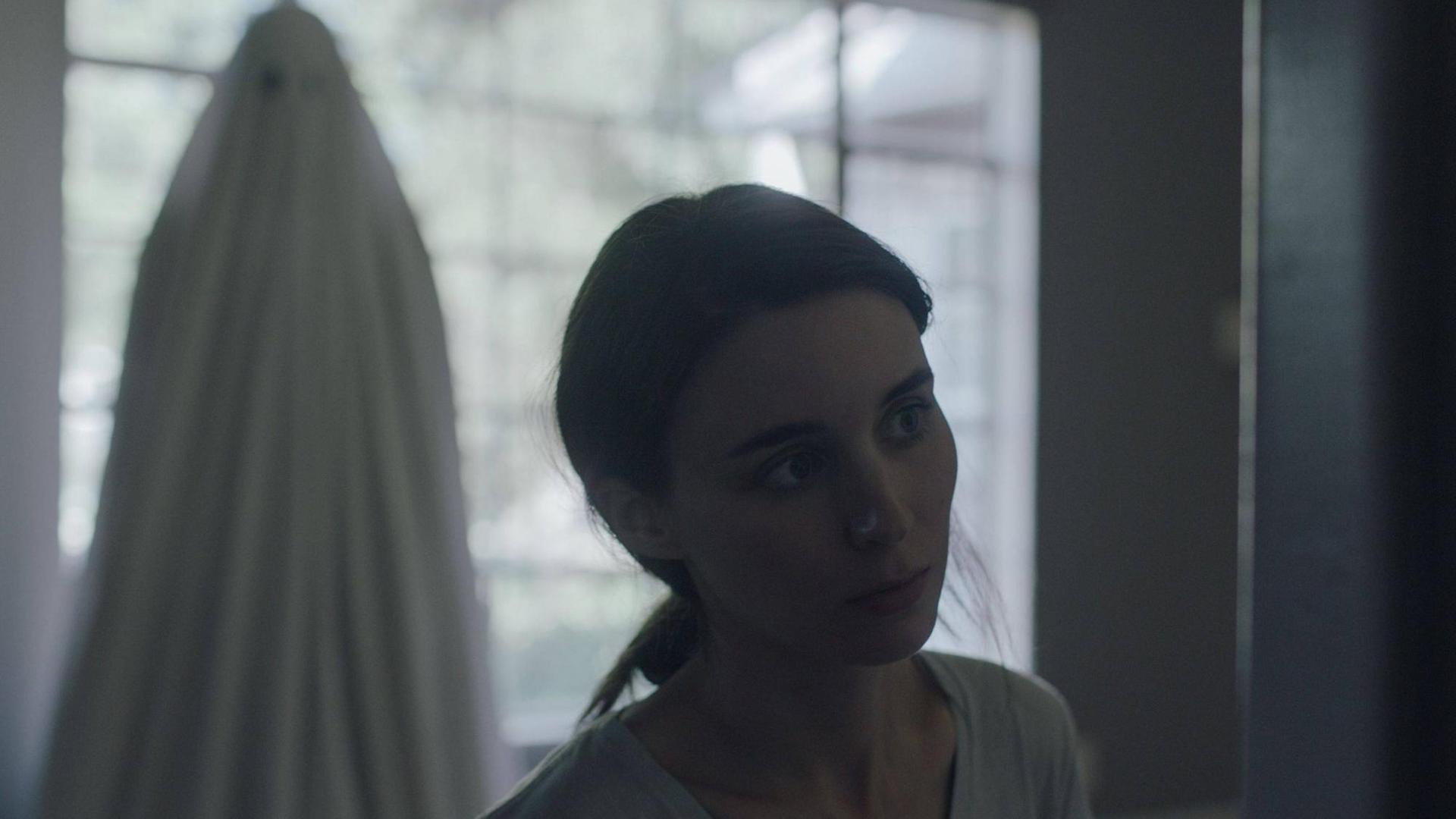 Szenenbild aus "A Ghost Story" - M (Rooney Mara) blickt traurig, hinter ihr der Geist von C (Casey Affleck) mit dem Bettlacken