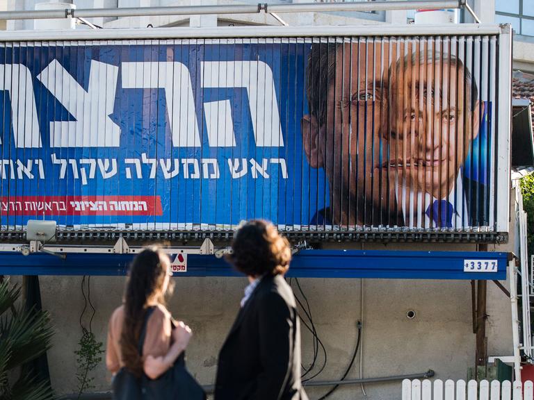 Am 17. März wählen die Israelis vorzeitig ein neues Parlament: Wahlberechtigte stehen vor einer Videowand, auf der Regierungschef Netanjahu und Oppositionsführer Herzog zu sehen sind.