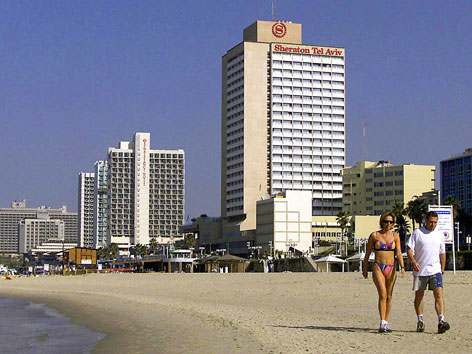 Zwei Personen gehen einen Strand entlang. Im Hintergrund sieht man mehrere Hochhäuser