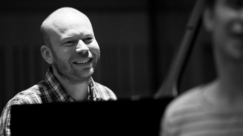 Auf dem s/w Bild ist hinter einem Flügel ist ein lachender kahlköpfiger Mann zu sehen. Er trägt Dreitagebart. Im Vordergrund rechts steht eine unkenntliche nur halb zu sehende Person