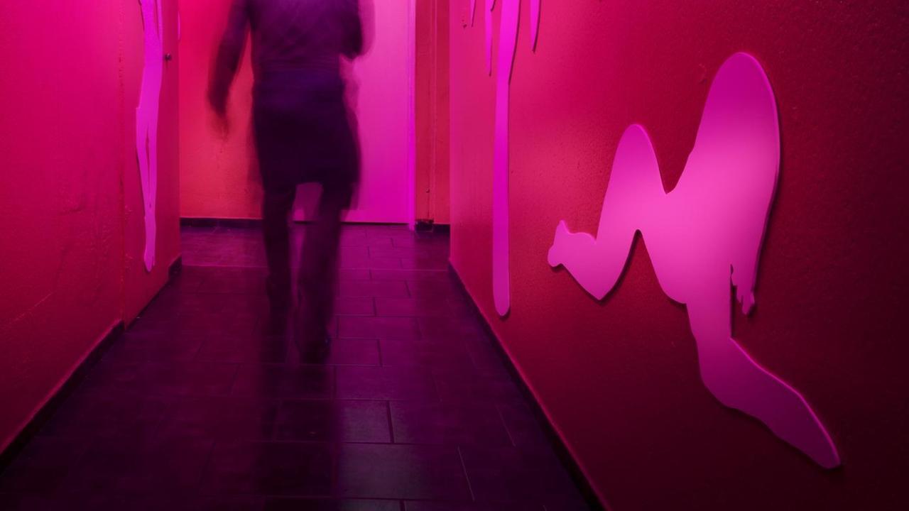Der Rücken einer Person ist in Bewegungsunschärfe auf einem rot beleuchteten Flur eines Bordells zu sehen. An den Wänden ist die Silhouette einer Fau zu erkennen.