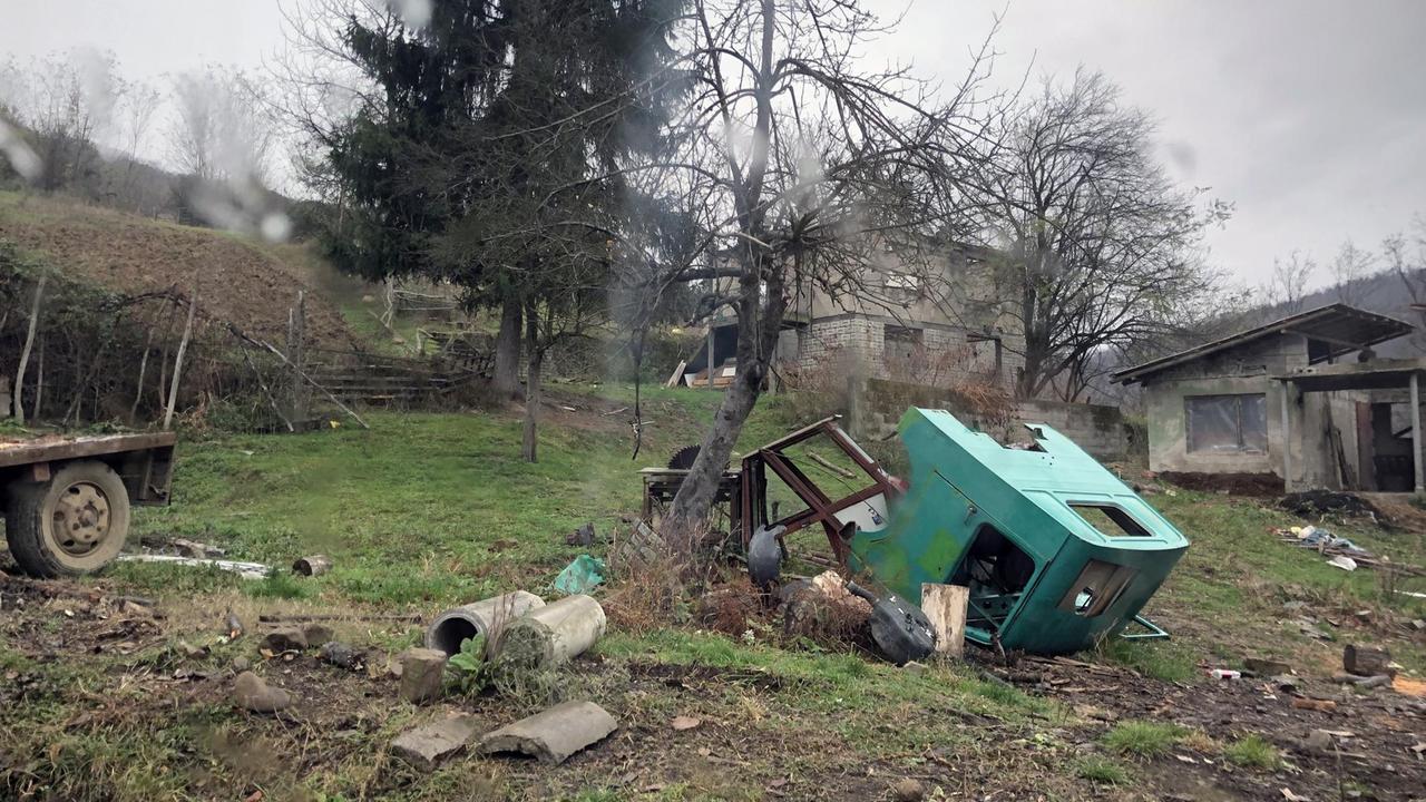 In einem bosnischen Dorf liegen alte Autoteile und ein Karren auf einer Wiese.