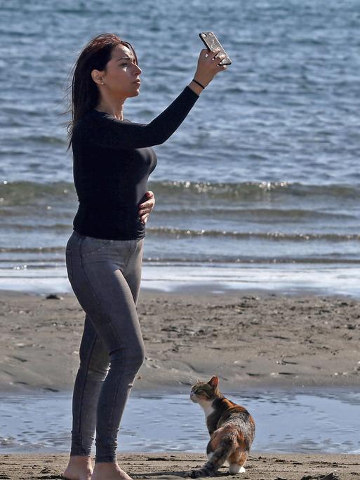 Eine Frau schießt am Strand von Limassol (Zypern) ein Selfie. Zu ihren Füßen sitzt ein braungescheckte Katze.