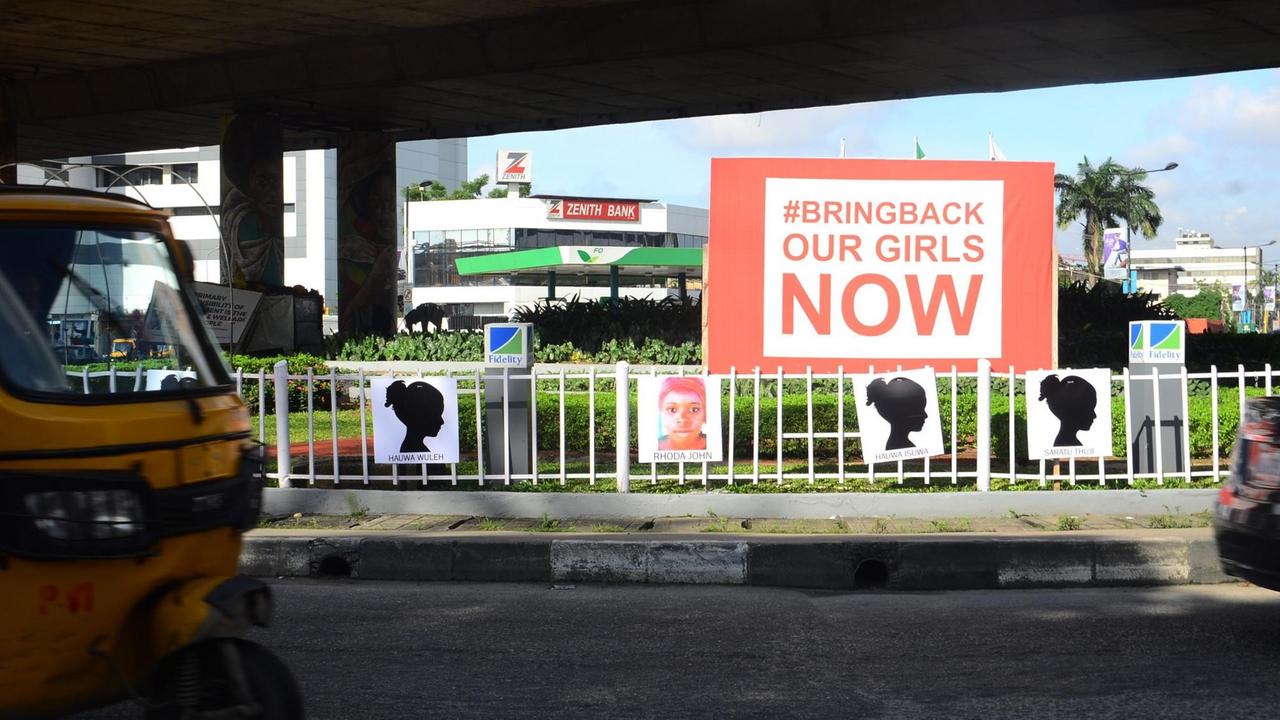An einer Straße sind Plakate mit den Namen der von Boko Haram entführten Mädchen aufgehängt. Darüber eine große Werbetafel mit der Aufschrift "Bring back our girls NOW".