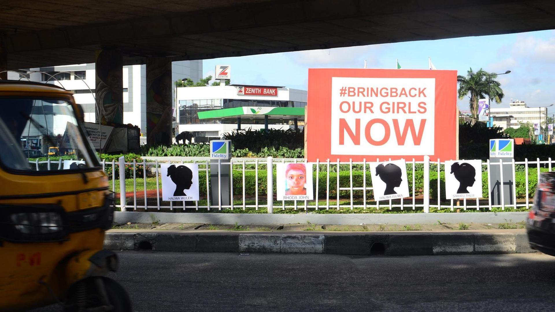 An einer Straße sind Plakate mit den Namen der von Boko Haram entführten Mädchen aufgehängt. Darüber eine große Werbetafel mit der Aufschrift "Bring back our girls NOW". An einer Straße sind Plakate mit den Namen der von Boko Haram entführten Mädchen aufgehängt. Darüber eine große Werbetafel mit der Aufschrift "Bring back our girls NOW".