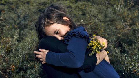 Ein Mädchen in einem blauen Kleid umarmt eine Frau, welche goldgelbe Blumen in der Hand hat.