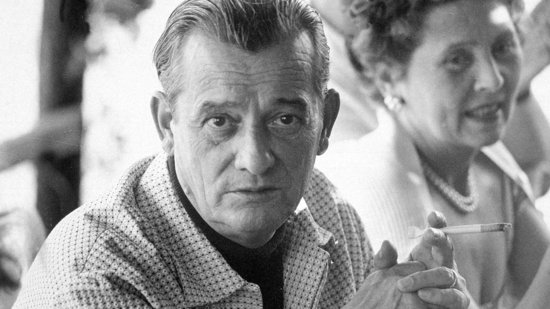 Der Schriftsteller Marcel Pagnol trägt ein gemustertes Hemd und hält eine Zigarette in der Hand. Das Foto wurde 1955 in Cannes aufgenommen.