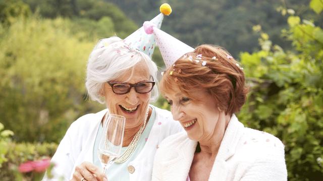 Zwei ältere Frauen mit Partyhütchen trinken auf einer Gartenparty Sekt und lachen.