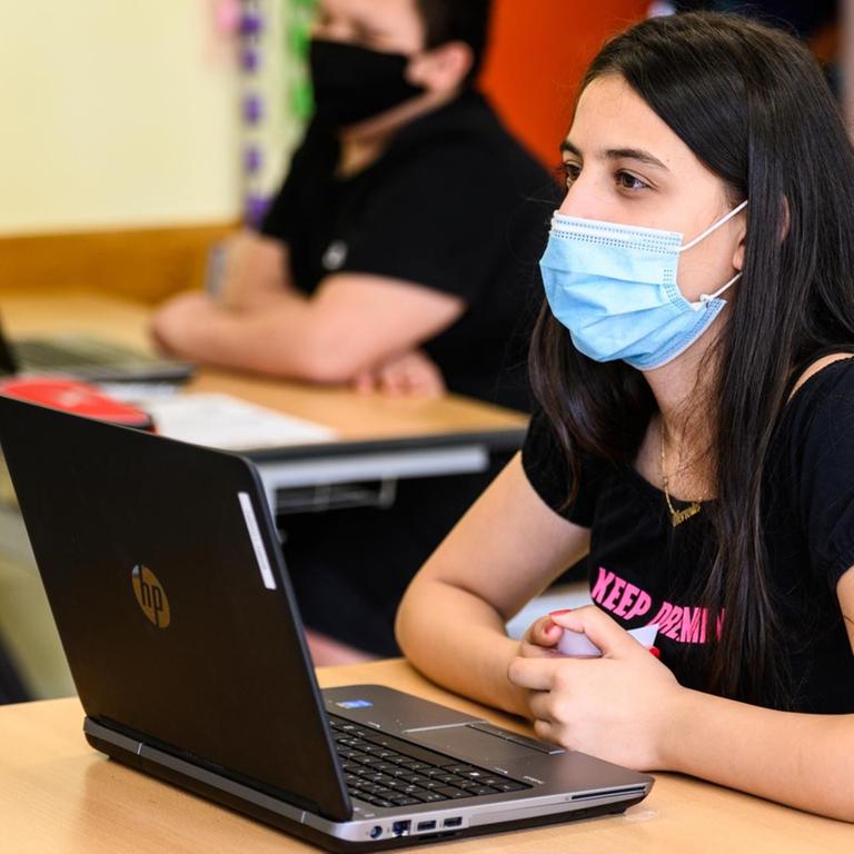 Schülerinnen und Schüler sitzen mit Maske und Laptop im Schulunterricht. 