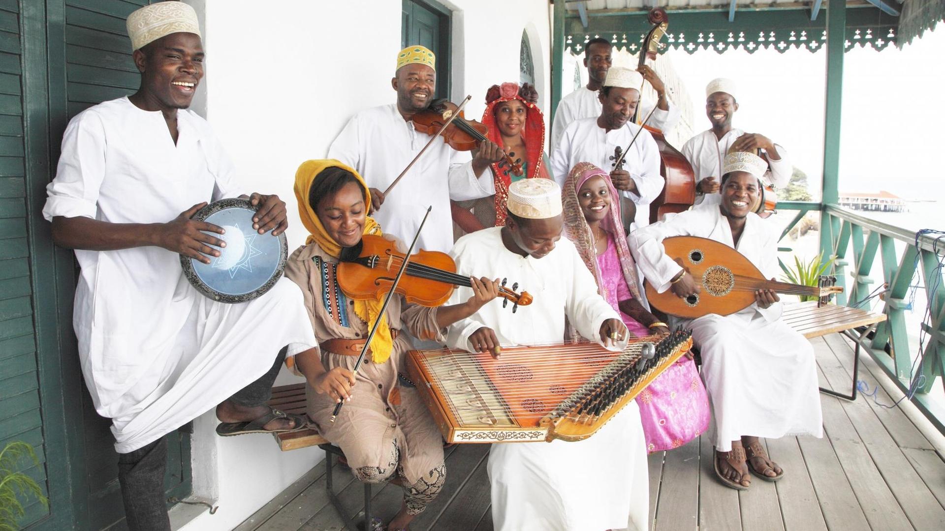 In traditioneller Kluft gekleidet steht eine Gruppe von farbigen Musikerinnen und Musikern lachend zusammen auf einer Holzveranda und spielen auf ihren Instrumenten.