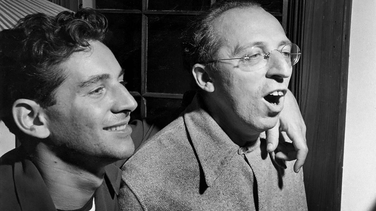 Schwarz-weiß Foto von 1945 mit den Komponisten und Dirigenten Leonard Bernstein und Aaron Copland. Sopland trägt eine runde, randlose Brille und singt offensichtlich