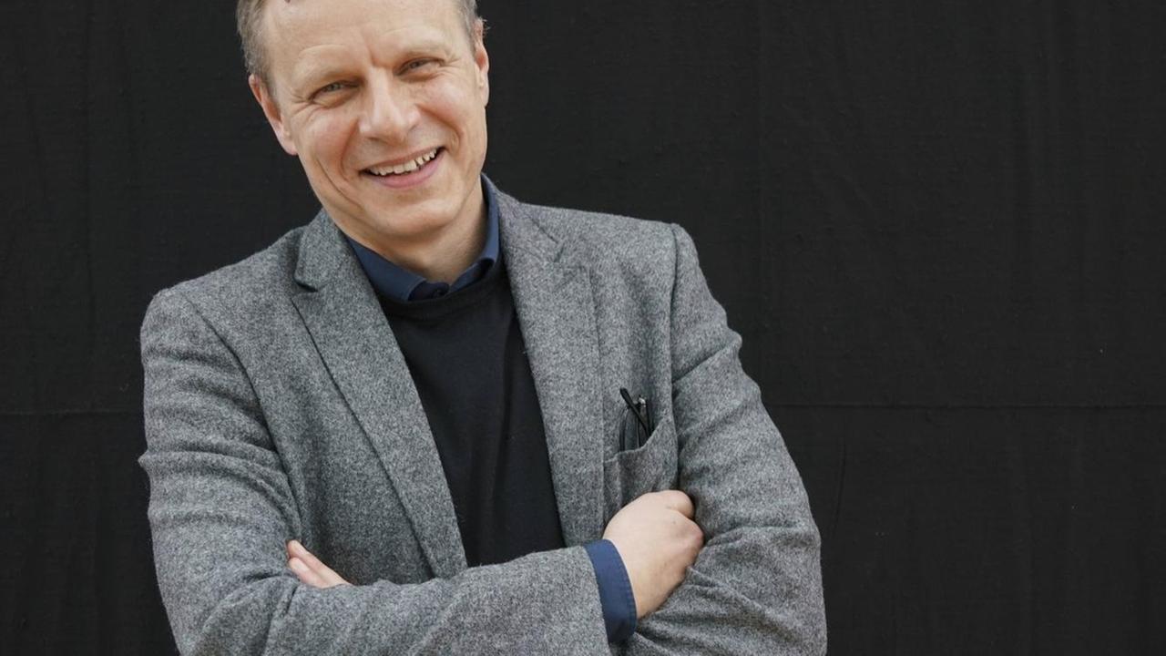Der Autor und Jurist steht vor einer schwarzen Wand, trägt ein graues Sacko und lacht in die Kamera.
