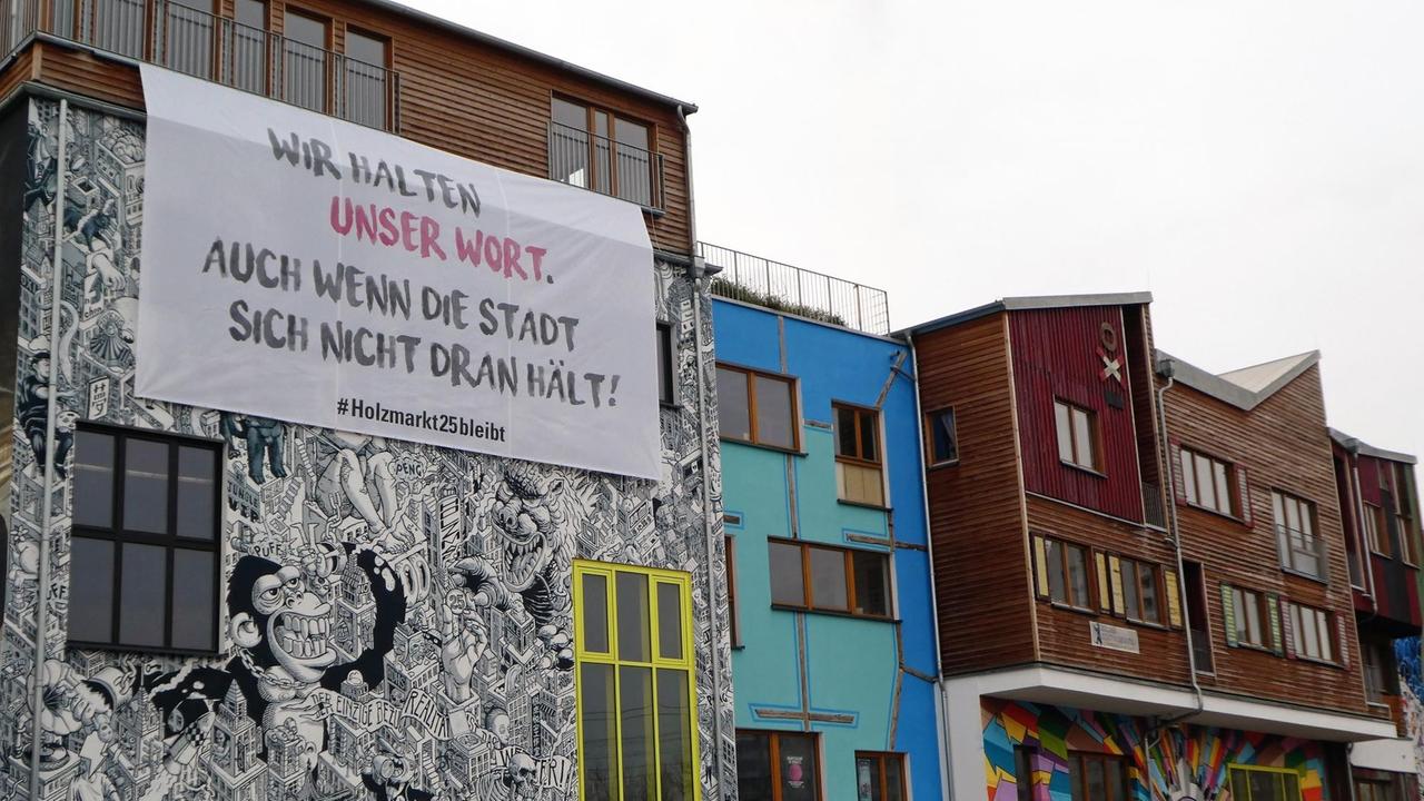 Blick auf die bemalten Hauswände des Berliner Holzmarktes. An einer Wand hängt ein Plakat, auf dem steht: Wir halten unser Wort, auch wenn die Stadt sich nicht dran hält! #Holzmarkt25 bleibt