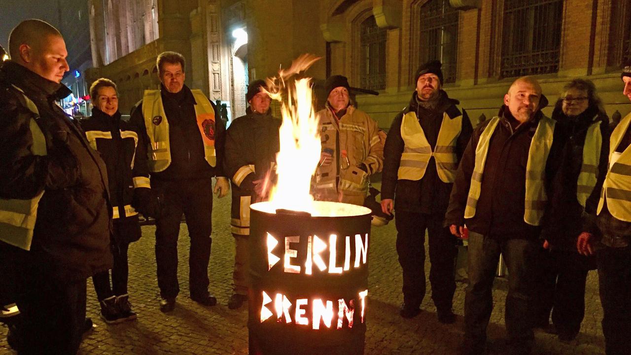 Neun Menschen stehen um eine Feuertonne, es ist dunkel. In die Tonne sind die Worte "Berlin brennt" eingestanzt, die nun durch die Tonne leuchten.