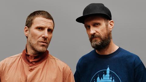 Zwei Männer blicken in die Kamera. Der linke Mann hat kurze Haare und trägt eine helle Jacke. Der rechte Mann trägt ein T-Shirt und eine Baseball-Kappe.
