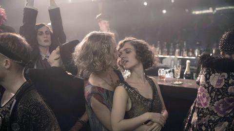 Partyszene aus der TV-Serie "Babylon Berlin" im Stil der 20er-Jahre: Eine Frau umarmt eine andere von hinten, ihre Gesichter sind einander zugewandt, darum mehrere Menschen, manche an einem Tresen, eine Frau reckt beide Arme in die Höhe.