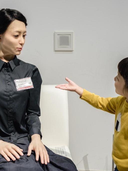 Ein junges Mädchen streckt einem weiblich aussehenden Roboter die Hand entgegen.