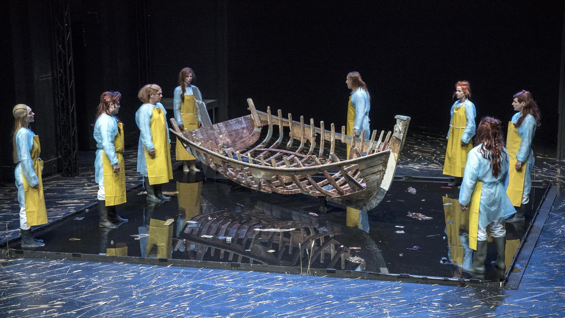 Ein Szenenbild des Theaterstücks "Boat Memory" von Roberto Ciulli zeigt das Gerippe eines Holzschiffes in der Mitte der Bühne. Daneben stehen links und rechts vereinzelt Menschen