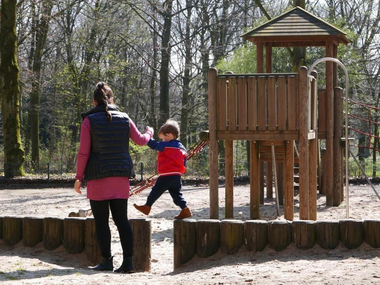 Eine Frau hält einen Jungen an der Hand während er auf einer Brüstung auf dem Spielplatz balanciert.