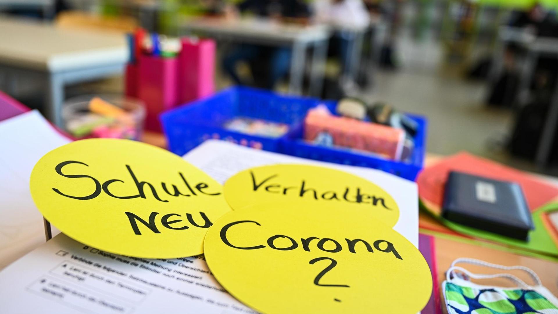 Auf dem Lehrerpult in einem Klassenzimmer liegen drei kreisrunde Papierschilder mit der Aufschrift: "Schule neu", "Verhalten" und "Corona ?", aufgenommen in der Achtalschule in Baienfurt in Baden-Württemberg im Mai 2020