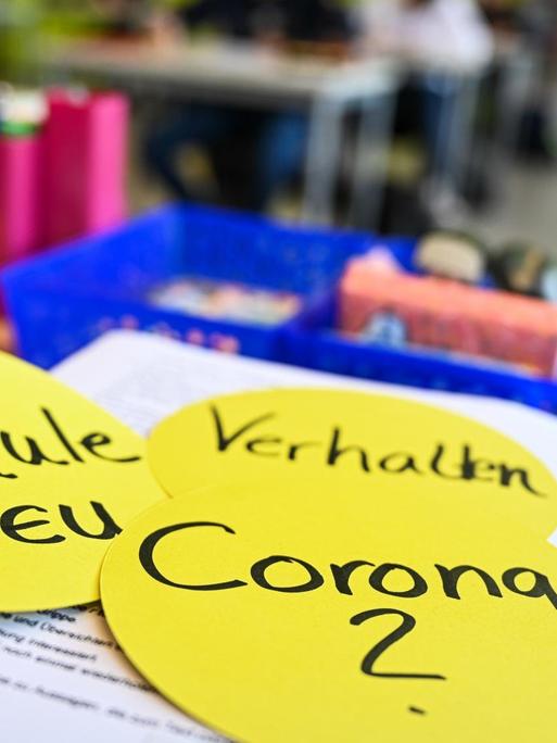 Auf dem Lehrerpult in einem Klassenzimmer liegen drei kreisrunde Papierschilder mit der Aufschrift: "Schule neu", "Verhalten" und "Corona ?", aufgenommen in der Achtalschule in Baienfurt in Baden-Württemberg im Mai 2020