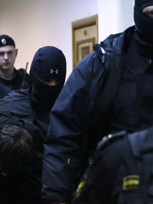 Mehrere Sicherheitskräfte in Uniform und teils vermummt begleiten einen Verdächtigen, der den Kopf gesenkt hält.