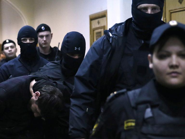 Mehrere Sicherheitskräfte in Uniform und teils vermummt begleiten einen Verdächtigen, der den Kopf gesenkt hält.