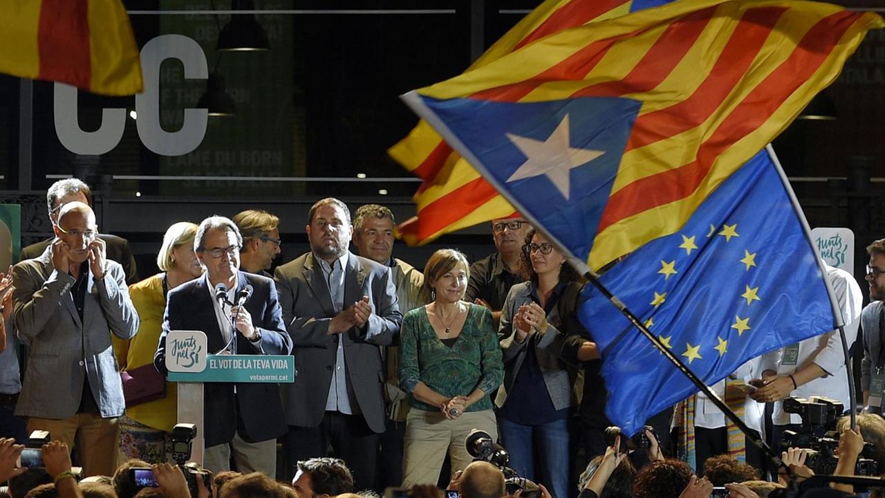 Auf einer Bühne stehen in der Nacht die Politiker, vor ihnen Wähler und Unterstützer. Am rechten Bildrand wehen eine Fahne Kataloniens und eine Europa-Fahne.