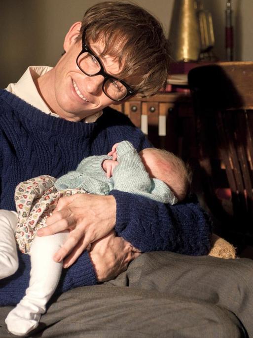 Eddie Redmayne als Stephen Hawking in einer Szene des Kinofilms "Die Entdeckung der Unendlichkeit" (2014). Das Foto zeigt den Hauptdarsteller als schwerbehinderten Physiker Stephen Hawking mit einen Baby auf dem Arm.