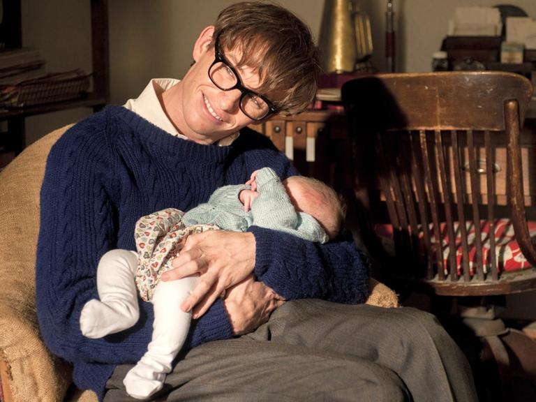 Eddie Redmayne als Stephen Hawking in einer Szene des Kinofilms "Die Entdeckung der Unendlichkeit" (2014). Das Foto zeigt den Hauptdarsteller als schwerbehinderten Physiker Stephen Hawking mit einen Baby auf dem Arm.