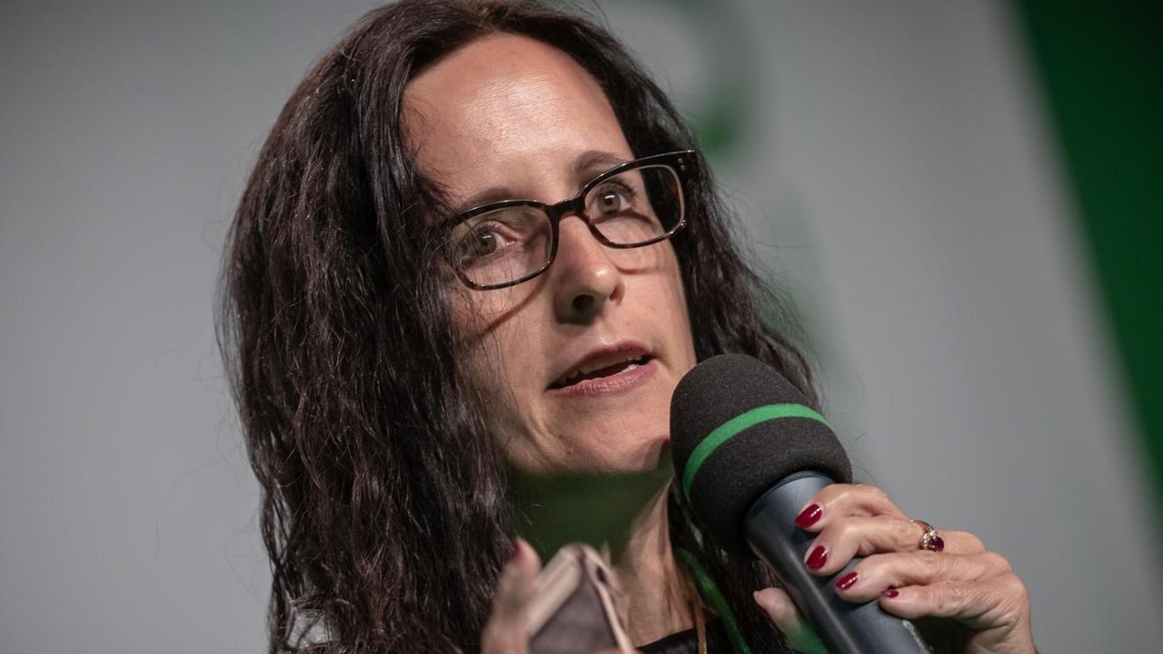 Porträtaufnahme Katharina Borcherts: eine Frau mit dunklen, langen Haaren und dunkel umrandeter Brille. In der Hand hält sie ein Mikrofon.
