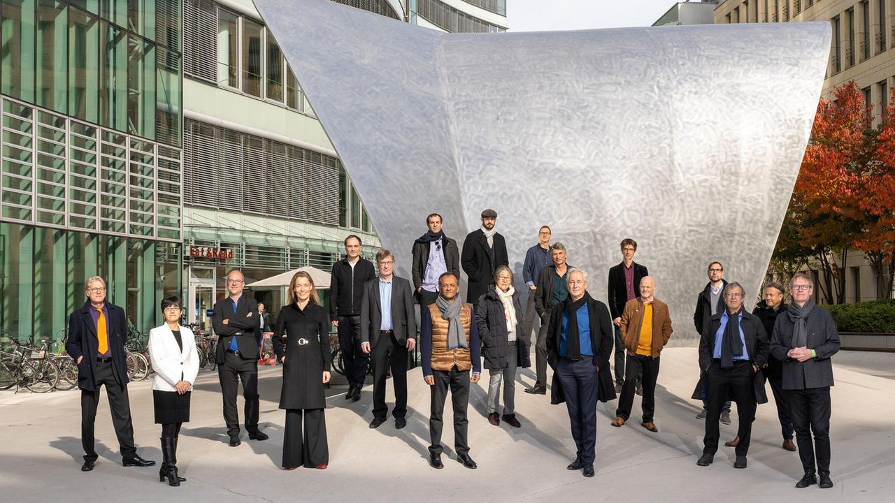 Eine Gruppe steht auf einem großzügigen modernen Platz mit einer großen gewölbten Metallskulptur, die sich wie ein volles Segel in die Höhe reckt.