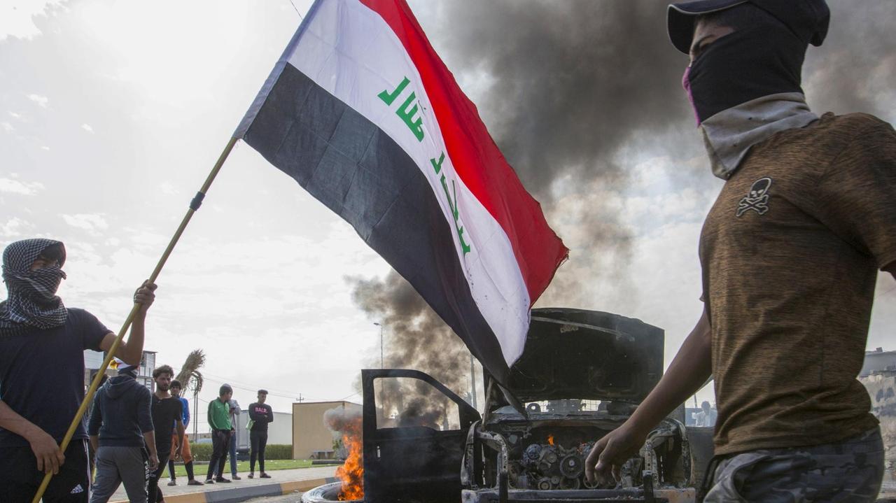 Irakische Demonstranten stehen neben einem ausgebrannten Autowrack.