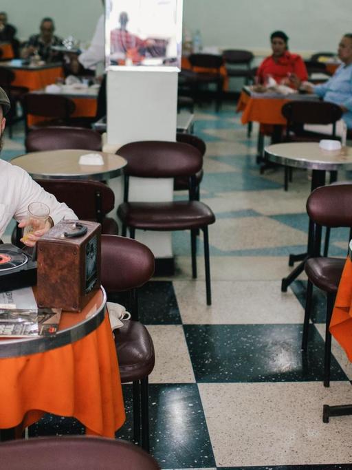 Ein Mann mit Schiebermütze, beigem Leinenhemd und Bart sitzt an einem kleinen runden Tisch, vor sich ein Kaffee. Mit der rechten Hand hebt er den Tonarm eines tragbaren Plattenspielers auf eine schwarze Vinyl-Single.
