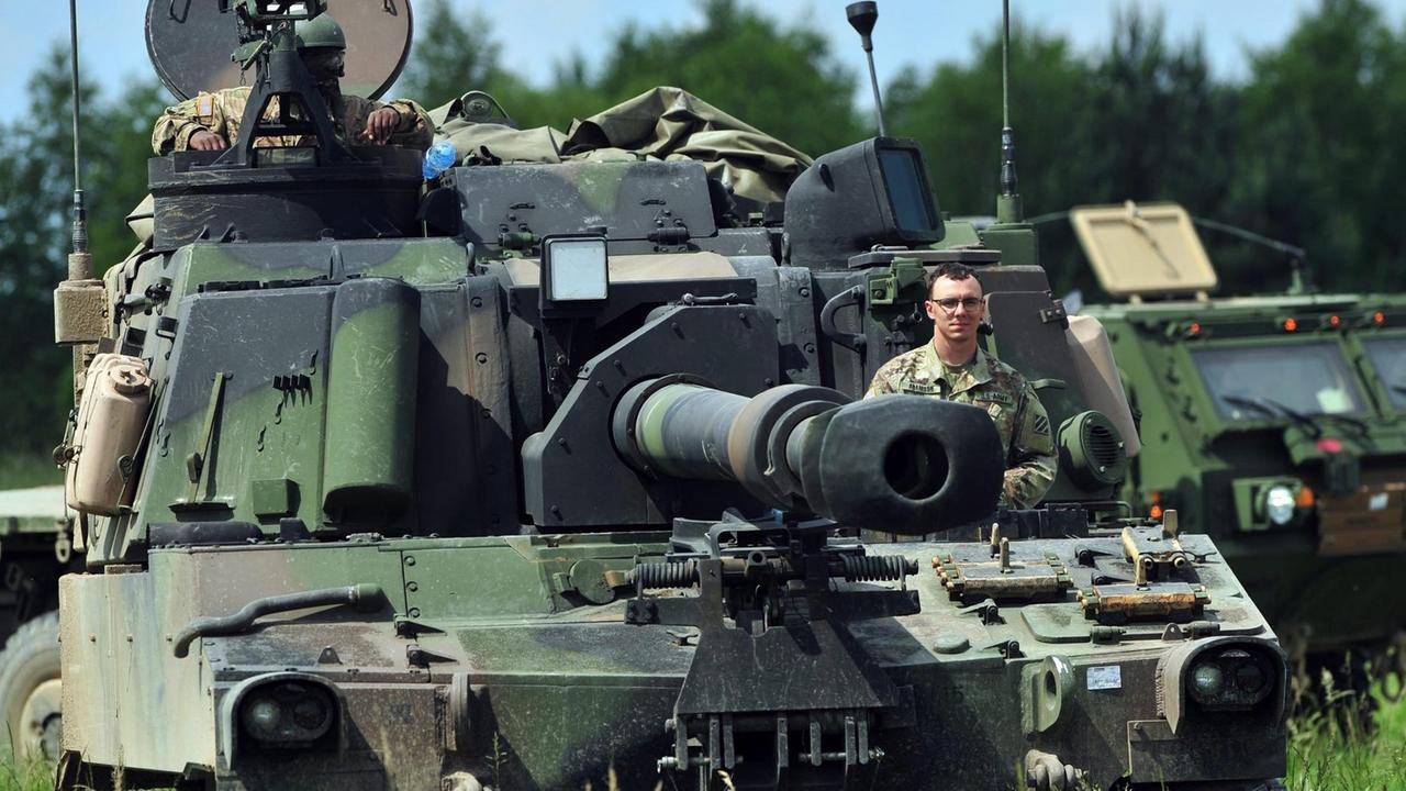 Man sieht einen Panzer mit zwei Soldaten, die aus den Luken scheuen. Dahinter ein Militärfahrzeug.