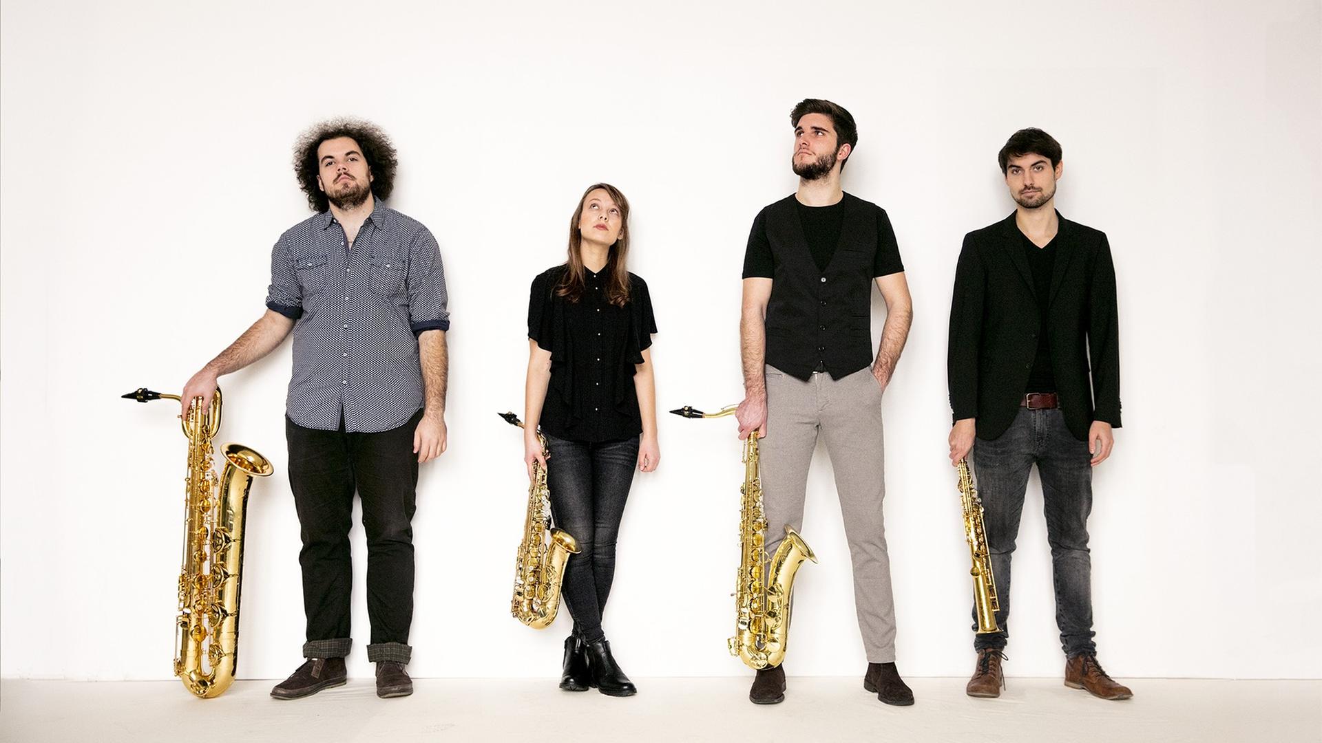 Drei Männer und eine Frau stehen überwiegend dunkel gekleidet mit vier unterschiedlich großen Saxophonen in der Hand vor einer weißen Wand.