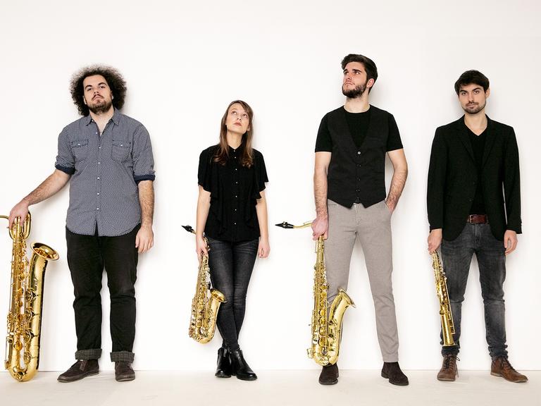 Drei Männer und eine Frau stehen überwiegend dunkel gekleidet mit vier unterschiedlich großen Saxophonen in der Hand vor einer weißen Wand.