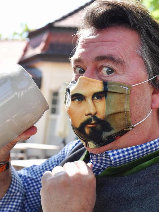 Ein Mann hebt einen Maßkrug, während er eine Gesichtsmaske mit einem Bild von König Ludwig trägt.