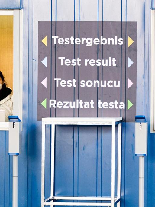 Zwei Personen in Schutzanzügen stehen an der größten Teststrasse Österreichs. Mehrsprachig ist "Testergebnis" zwischen den Fenstern geschrieben.