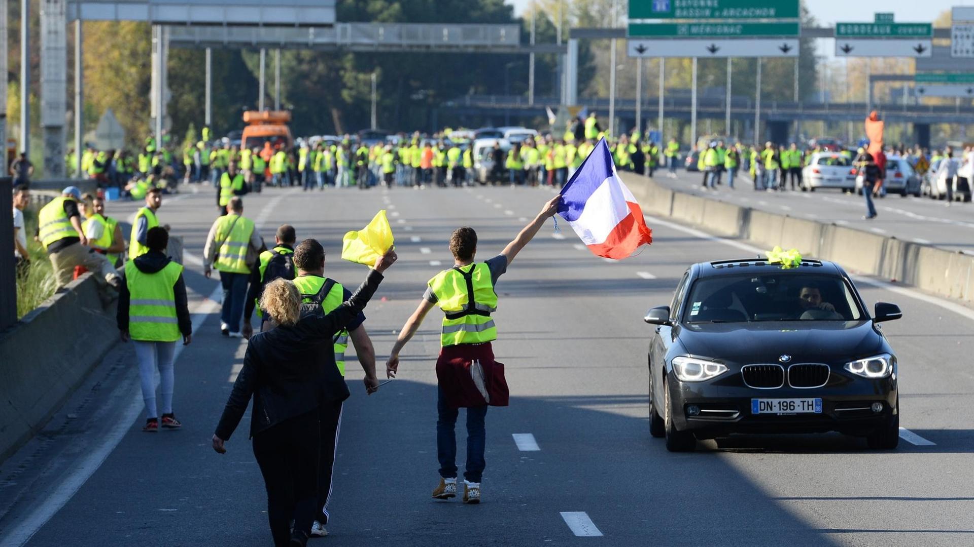 Demonstranten in gelben Warnwesten laufen auf einer mehrspurigen Straße, ein Mann schwenkt die französische Flagge.