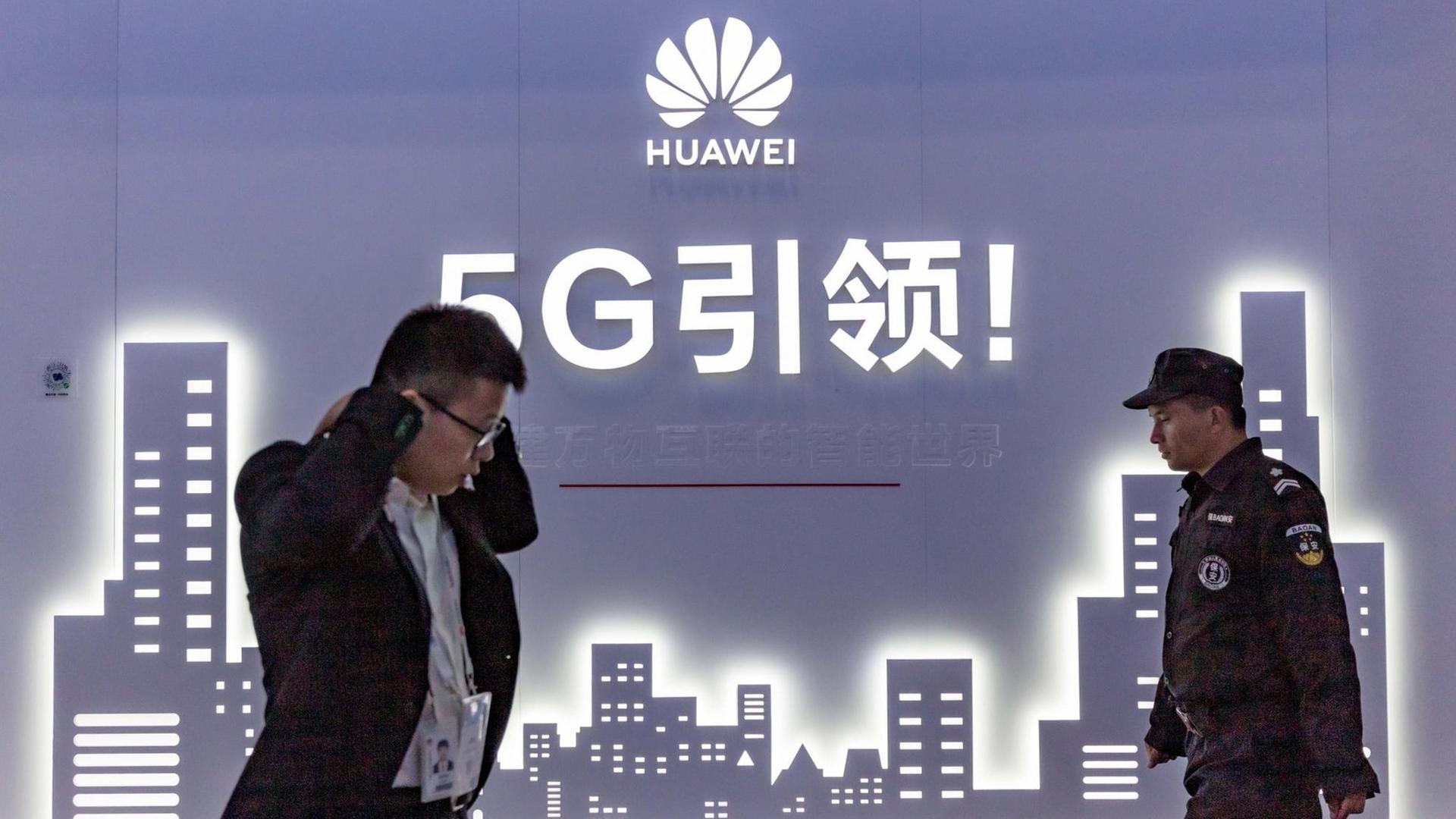 An der Wand eine blaue Grafik mit Hochhäusern, darüber stehen mit weißer Schrift Huawei und 5G sowie chinesische Schriftzeichen. Vor der Wand links ein Mann, der im Laufen telefoniert, von rechts kommt ein Uniformierter.