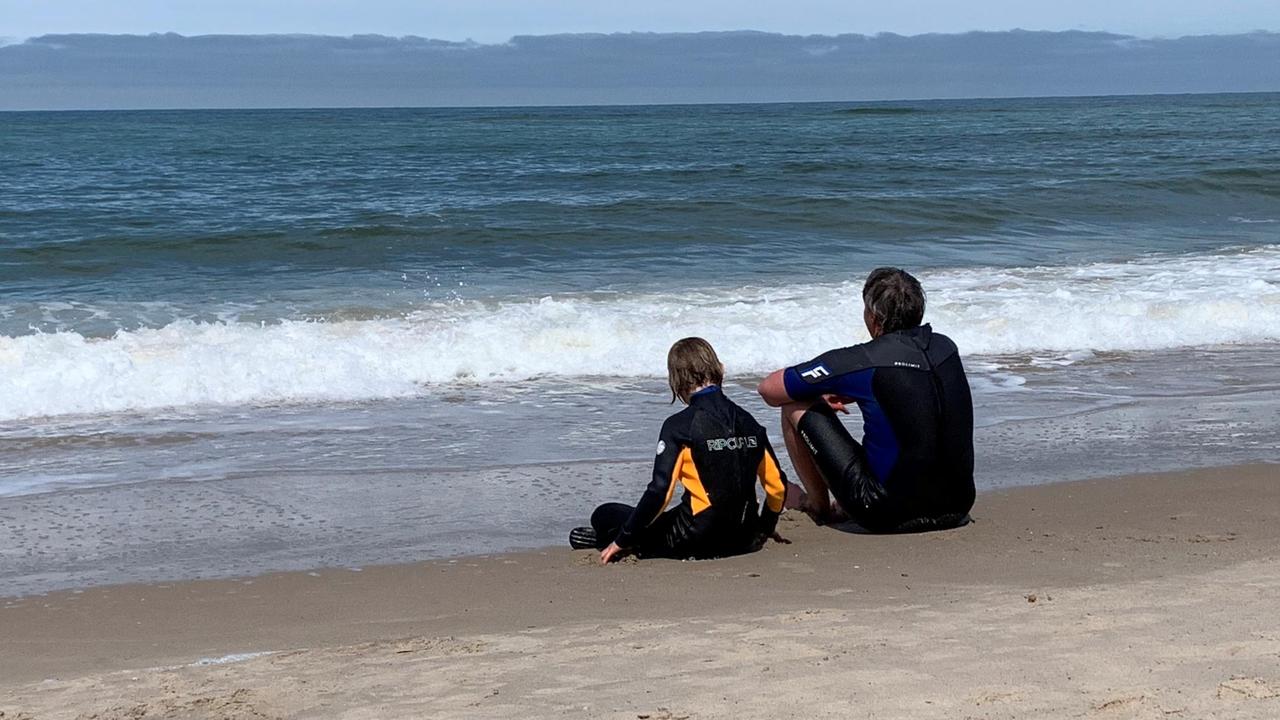 Mutter und Tochter tragen Neoprenanzüge und sitzen am Strand. Sie blicken aufs Meer.